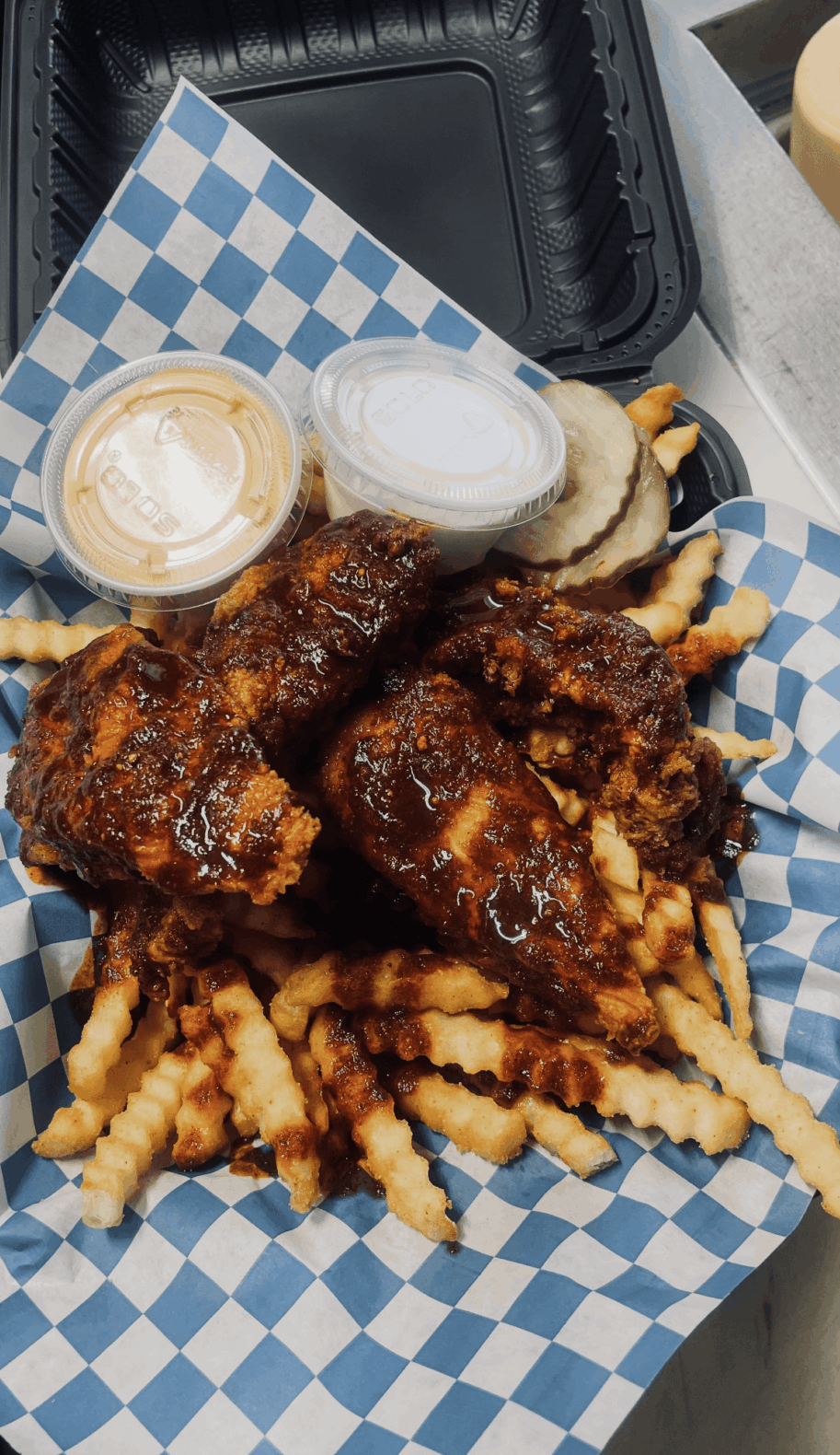 Basket with fried chicken, French fries, pickles, and dipping sauces on blue checkered paper.