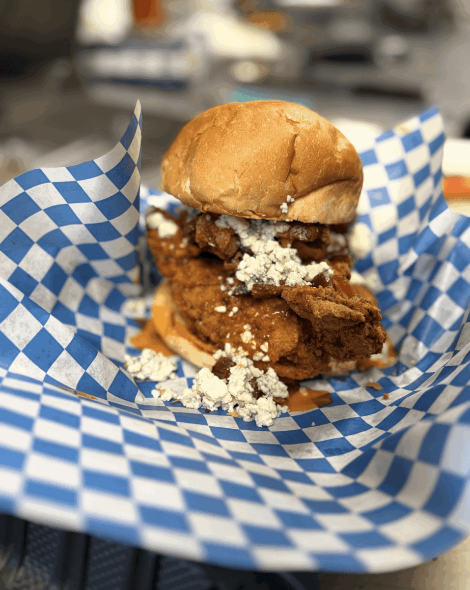Fried chicken sandwich with coleslaw in a bun on blue checkered paper, with a blurred background.