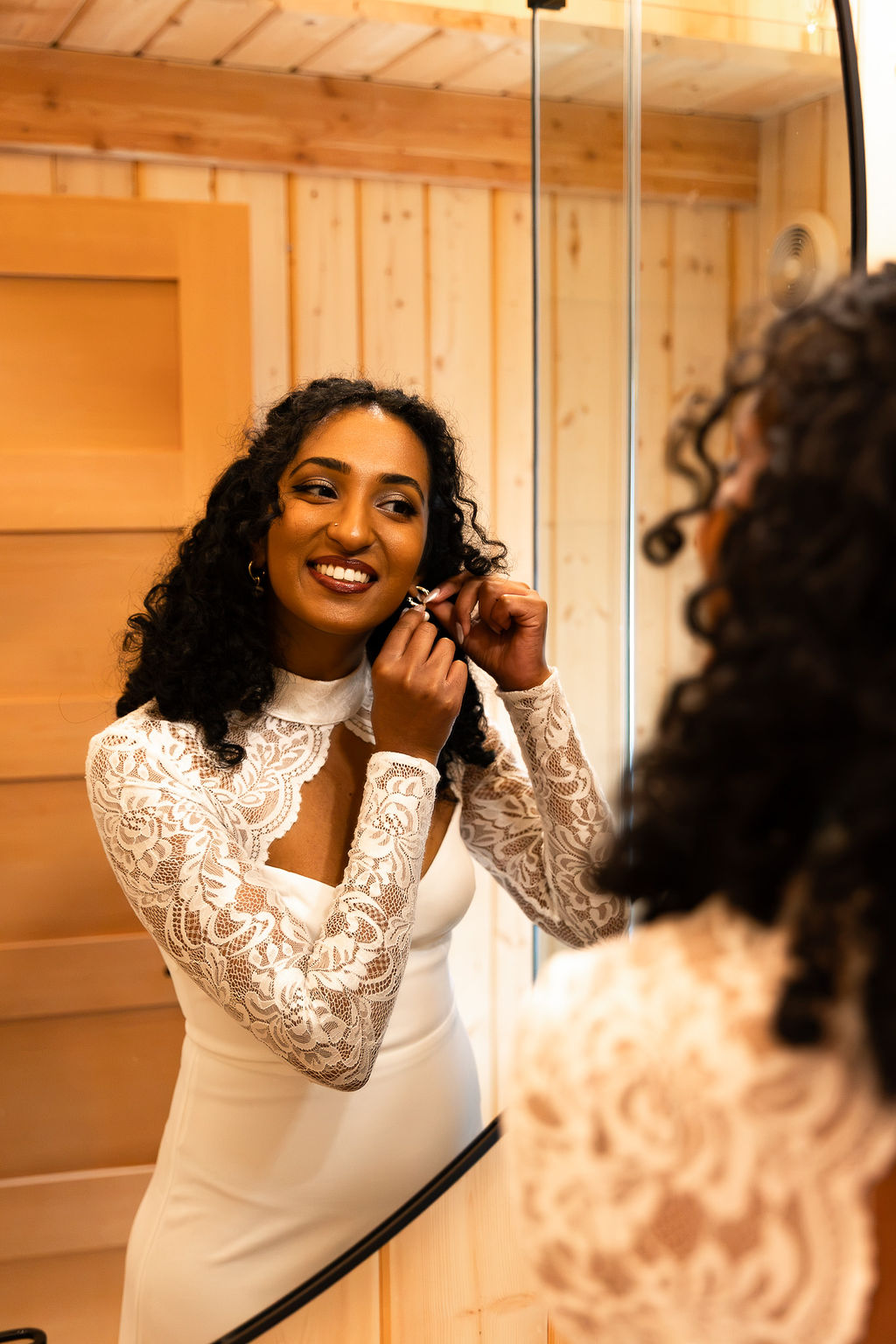 Woman with curly hair in a white lace dress smiling while adjusting her earring, reflected in a mirror.