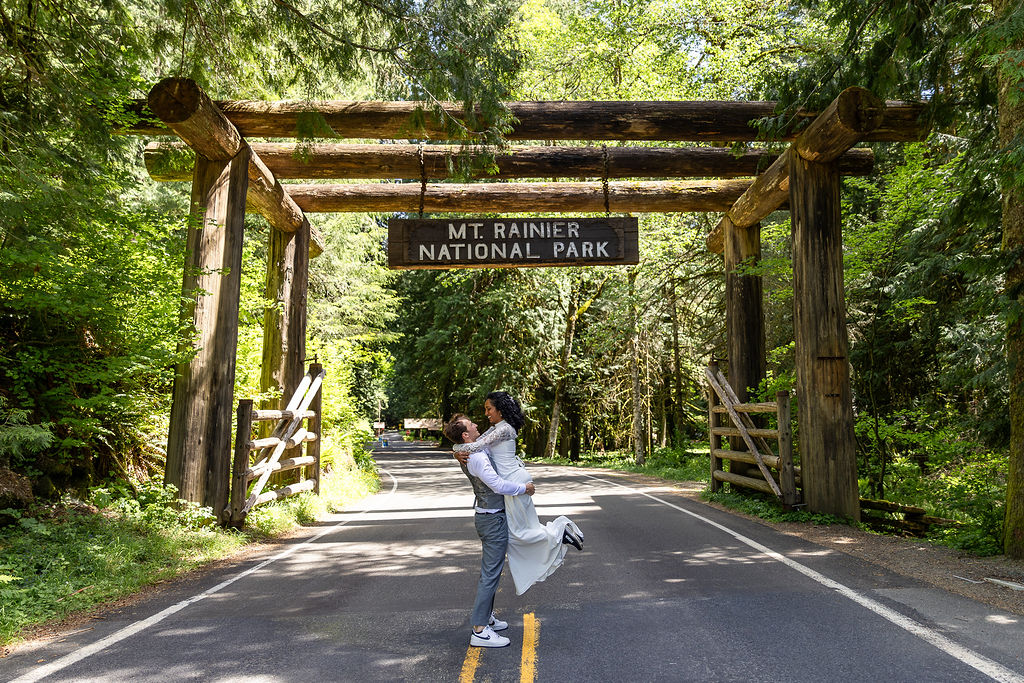 Person standing on a road under a wooden archway at Mount Rainier National Park, surrounded by trees.