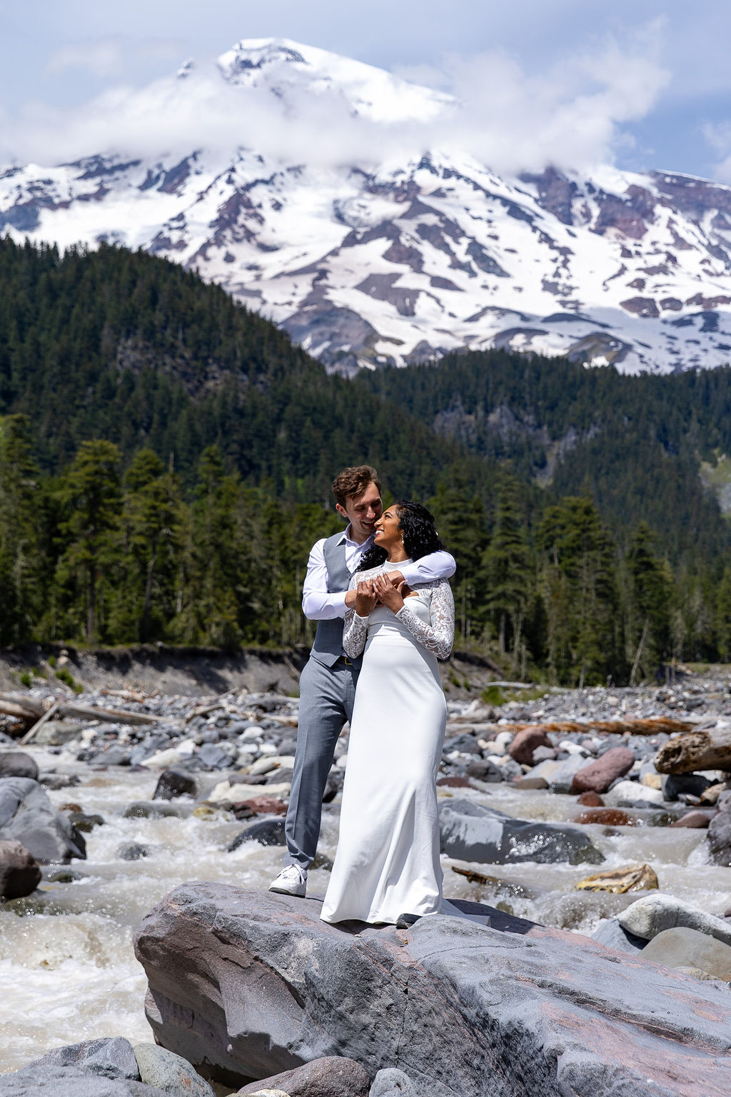 A couple in wedding attire stands on rocks in a river with snow-capped mountains and forest in the background.