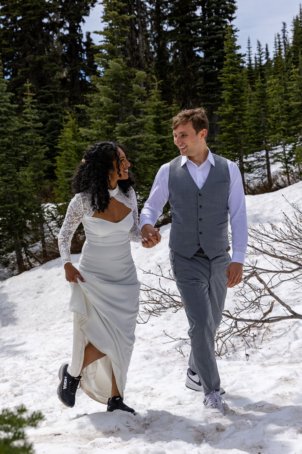 A couple holding hands walking in snow with trees in background, woman in white dress, man in gray vest and pants.
