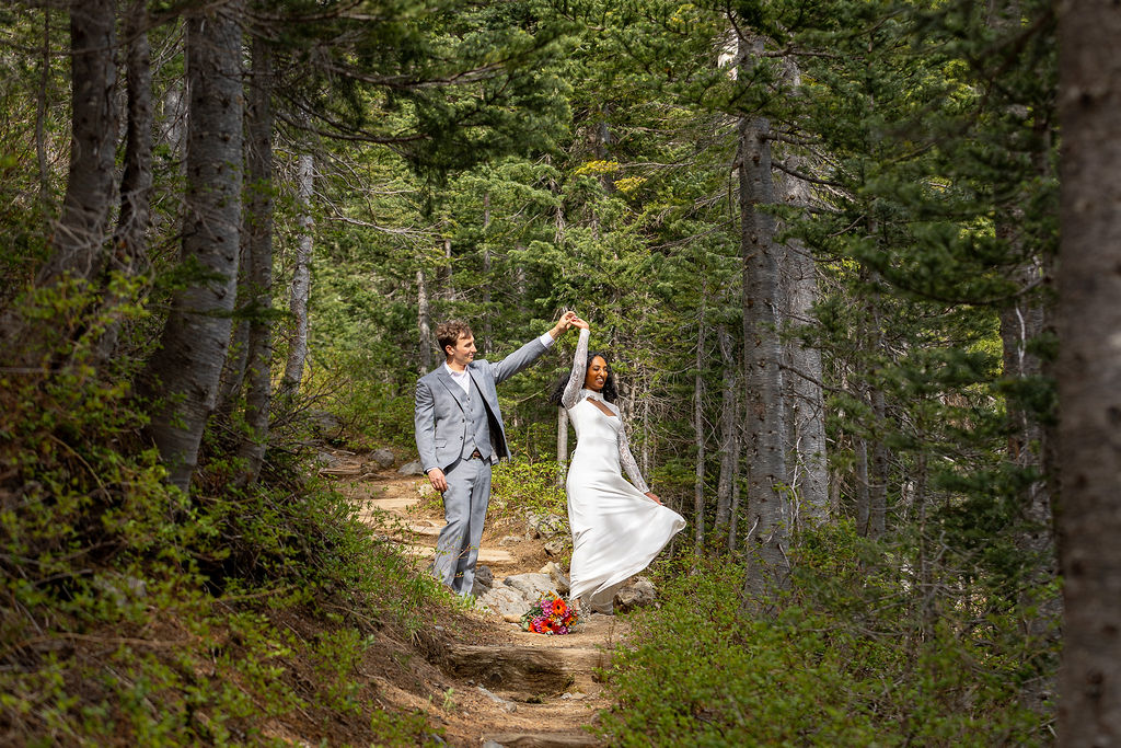 A couple dancing in a forest, with trees and greenery around them, the woman in a white dress and the man in a suit.