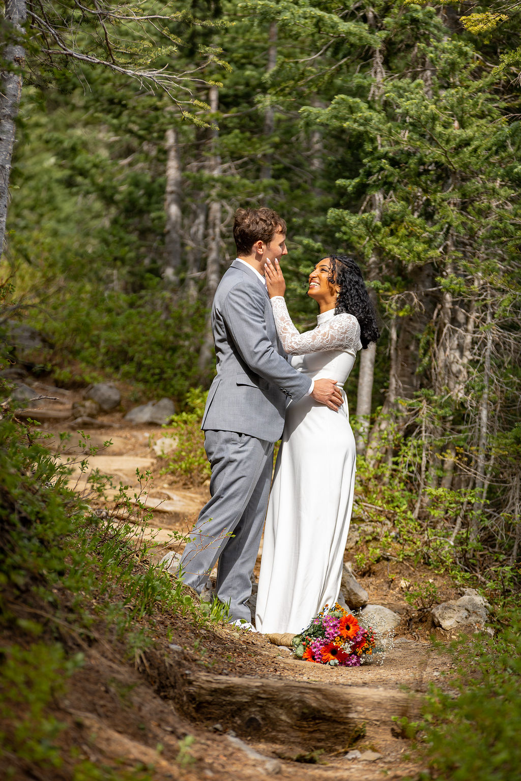 A couple in wedding attire standing on a forest path, embracing and looking at each other, with a bouquet on the ground.