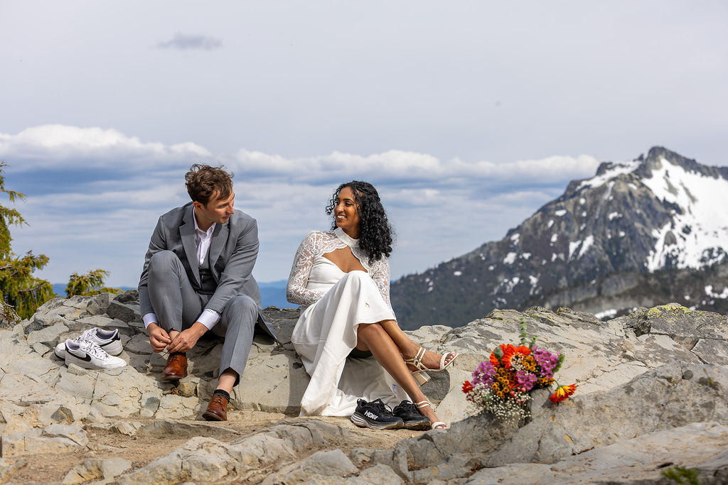 Two people sitting on rocks outdoors with a mountain and cloudy sky in background, flowers nearby, one person in a suit, the other in a white dress.