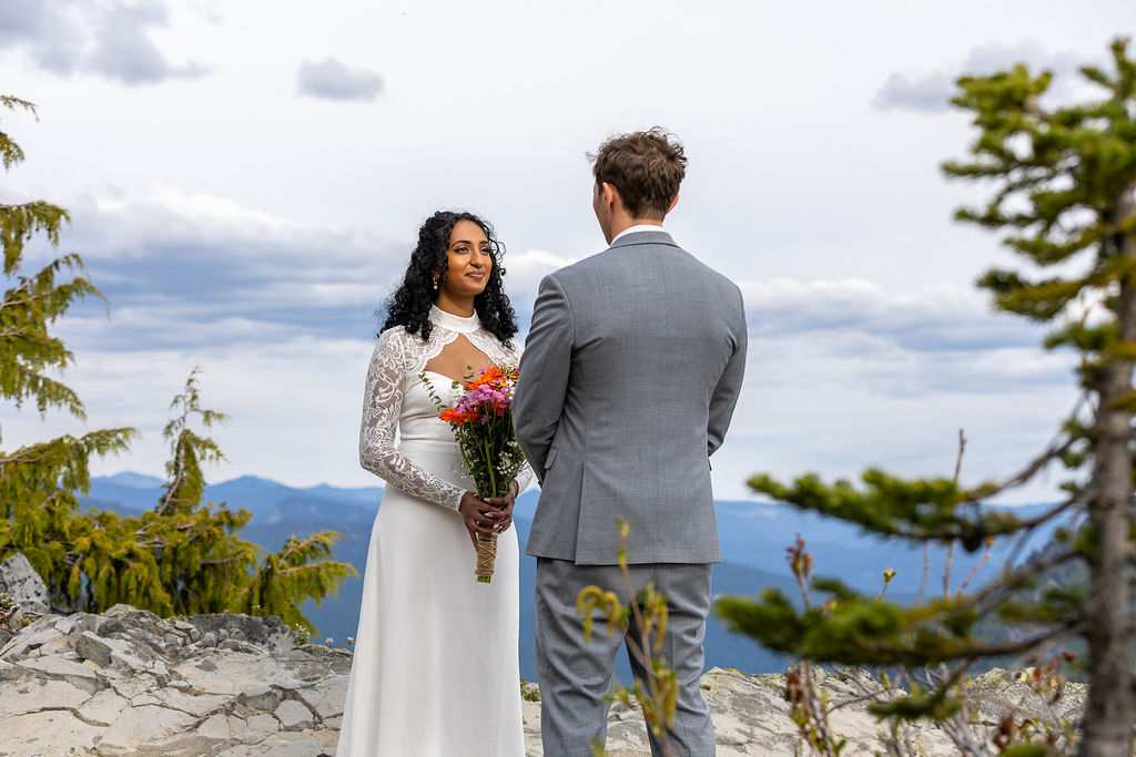 A couple stands outdoors on a rocky area with trees and mountains in the background, exchanging vows or a moment of connection.