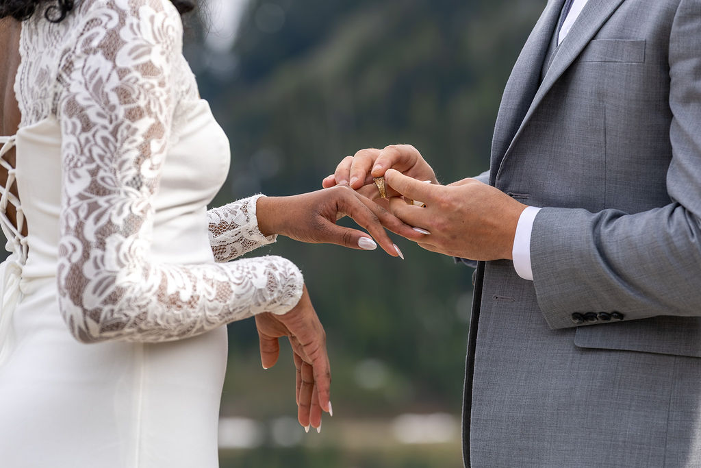 A couple exchanging rings outdoors, with a blurred natural background, focusing on their hands and wedding rings.