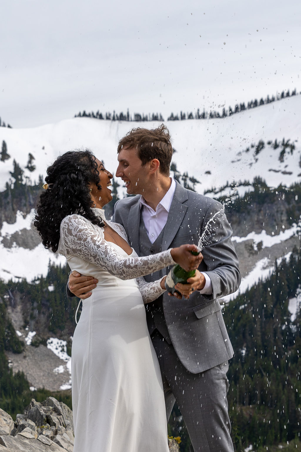 A couple in wedding attire celebrating outdoors in a snowy mountain landscape, holding a bottle and spraying champagne.