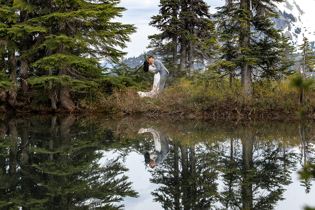 Person standing on a grassy bank near a calm lake with trees and mountains in the background, reflected in the water.