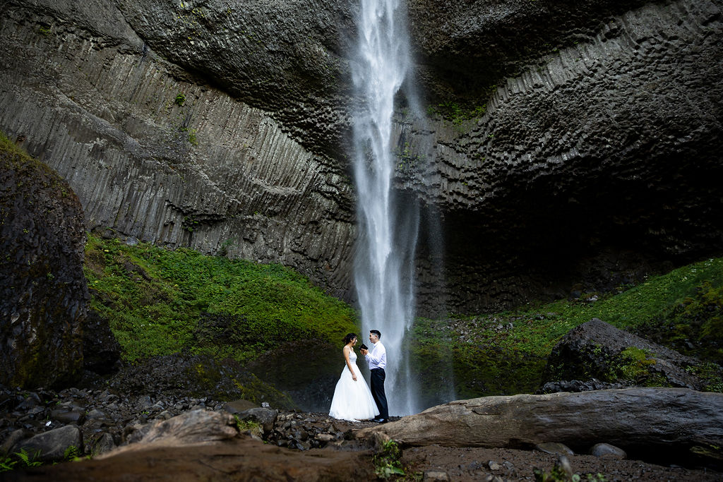 A couple stands under a tall waterfall in a rocky, green landscape, with water cascading down from above.