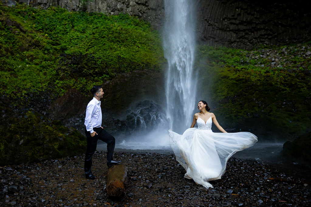 A woman in a white dress and a man in a white shirt and dark pants stand near a waterfall with lush green mossy rocks behind them.