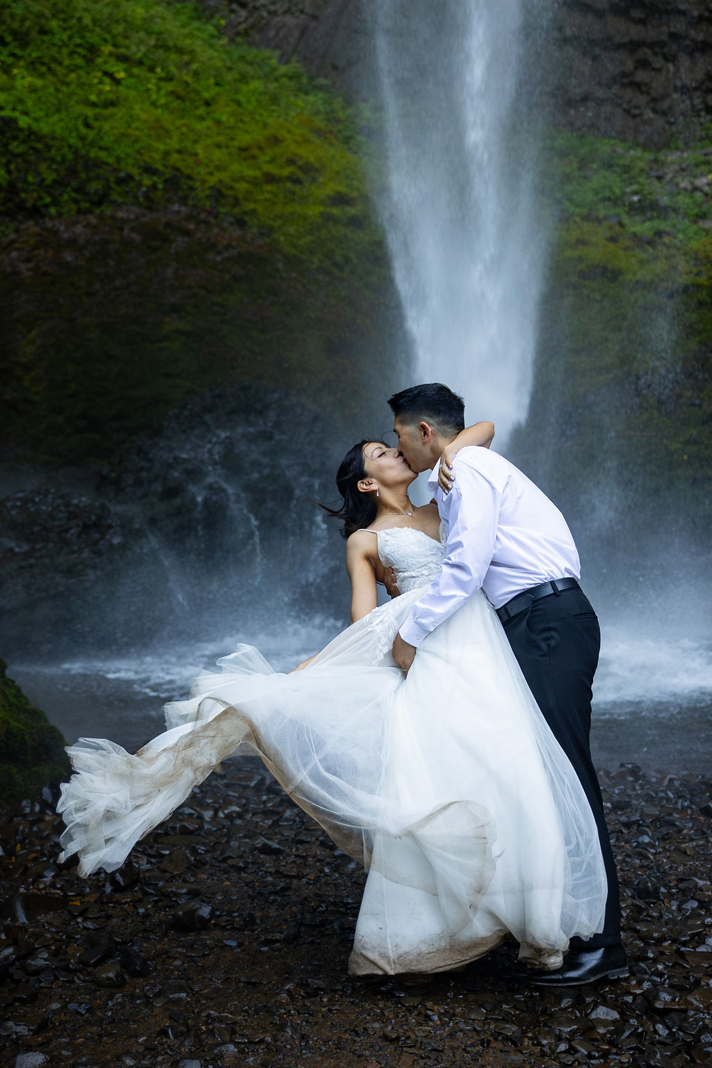 Couple in wedding attire embracing in front of a waterfall, with water cascading behind them.