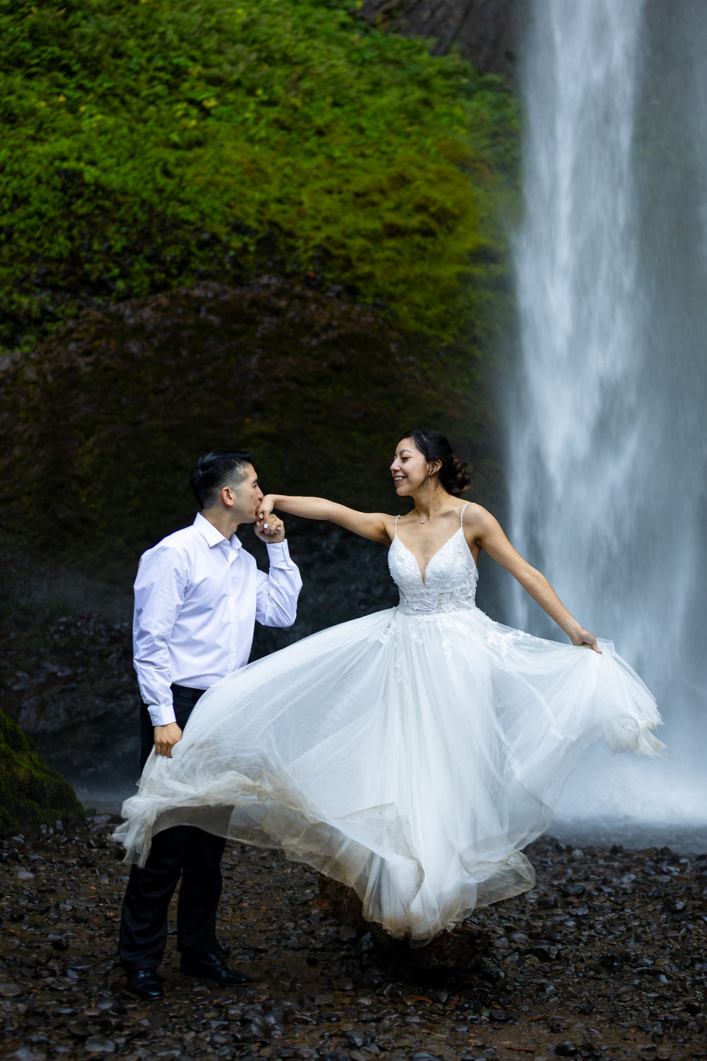 A couple in wedding attire stands near a waterfall, with the bride's dress flowing and the groom holding her hand.