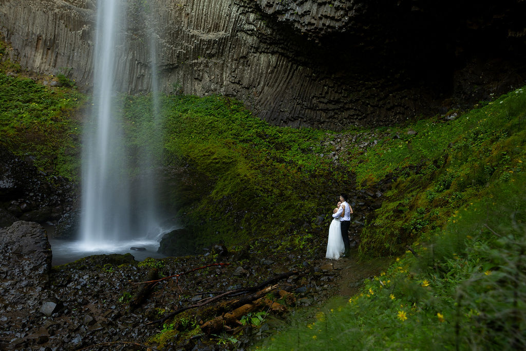 A person in a white dress stands near a waterfall in a lush green landscape.