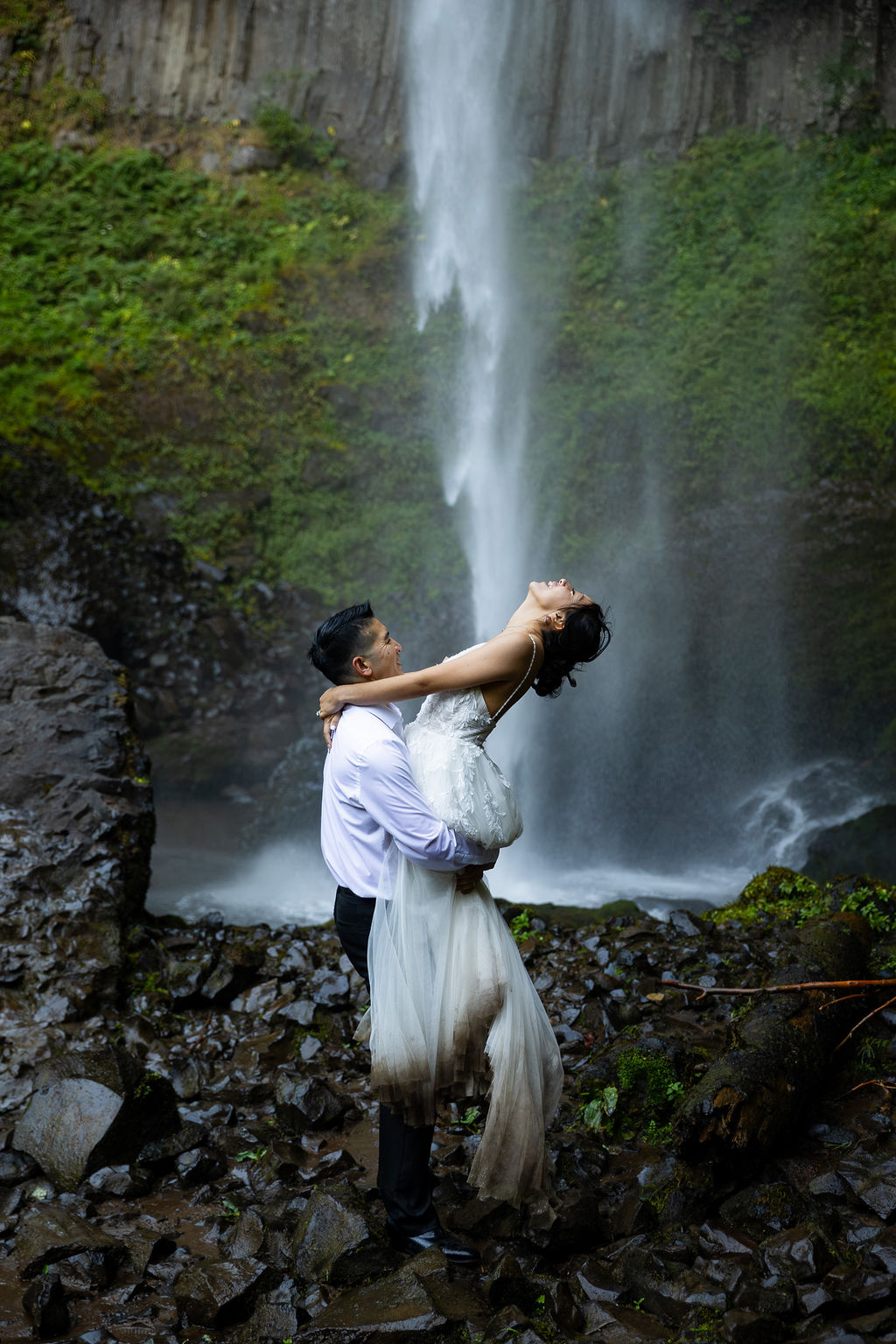 A couple stands near a waterfall, with the man lifting the woman in front of the cascading water.