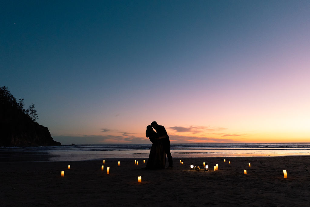 Silhouette of a couple kissing on a beach at sunset, with small lights on the sand and a calm ocean background.