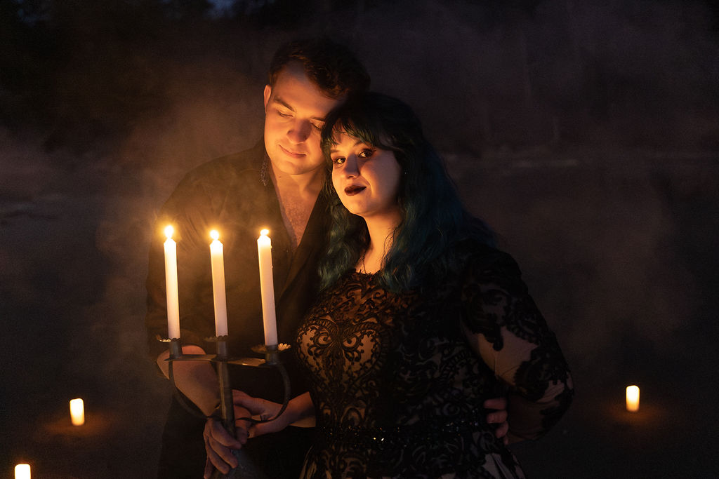 Two people holding a candelabrum with three lit candles, surrounded by a dark, smoky background with small candles, one on each side.