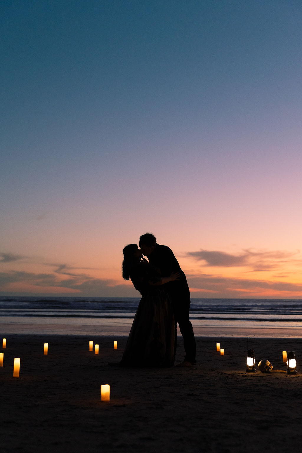 Silhouette of a couple embracing on a beach at sunset, with candles on the sand and a colorful sky.