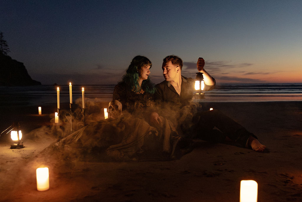Two people sitting on a beach at sunset, surrounded by candles and lanterns, with a body of water in the background.