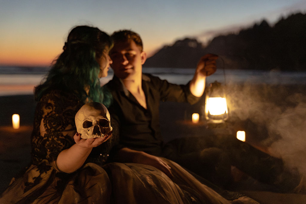 Two people sitting on a beach at sunset, holding a lantern and a skull, with candles around them, foggy atmosphere.