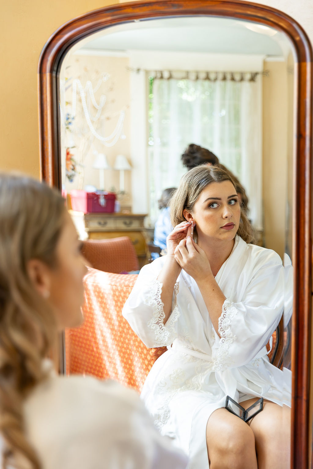 Young woman in white robe looking at herself in mirror, adjusting earring, in a bright room with curtains and furniture