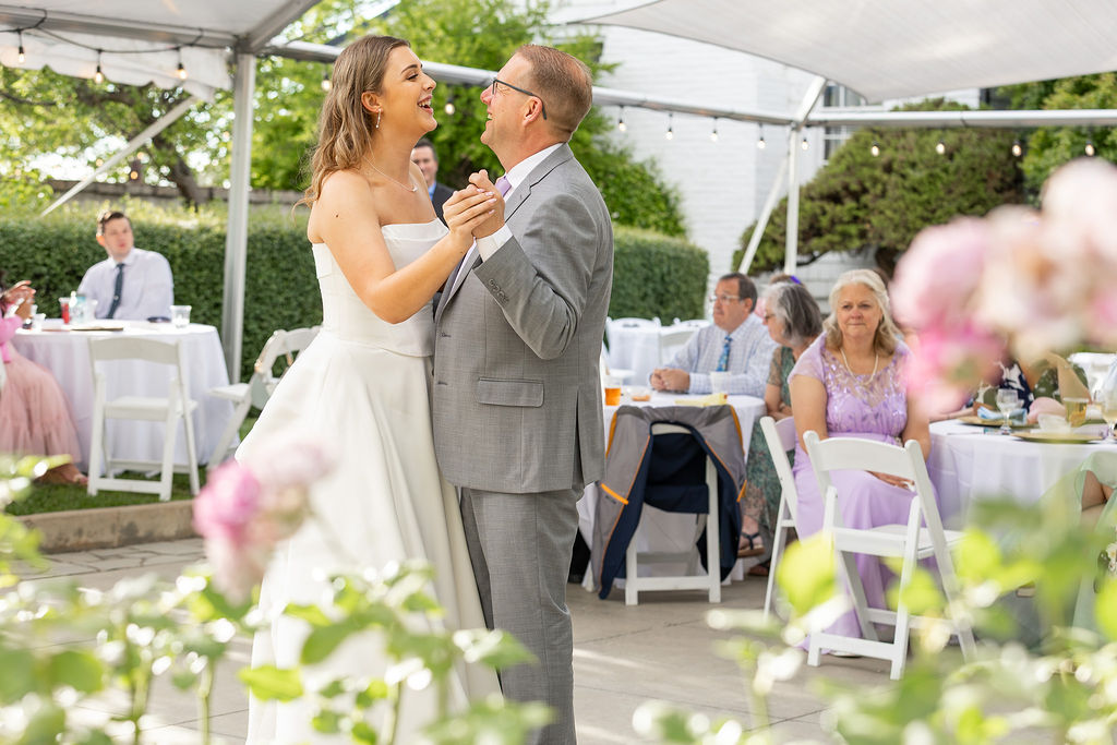 A bride and groom dance outdoors under a tent, surrounded by guests at tables with pink and white decorations.