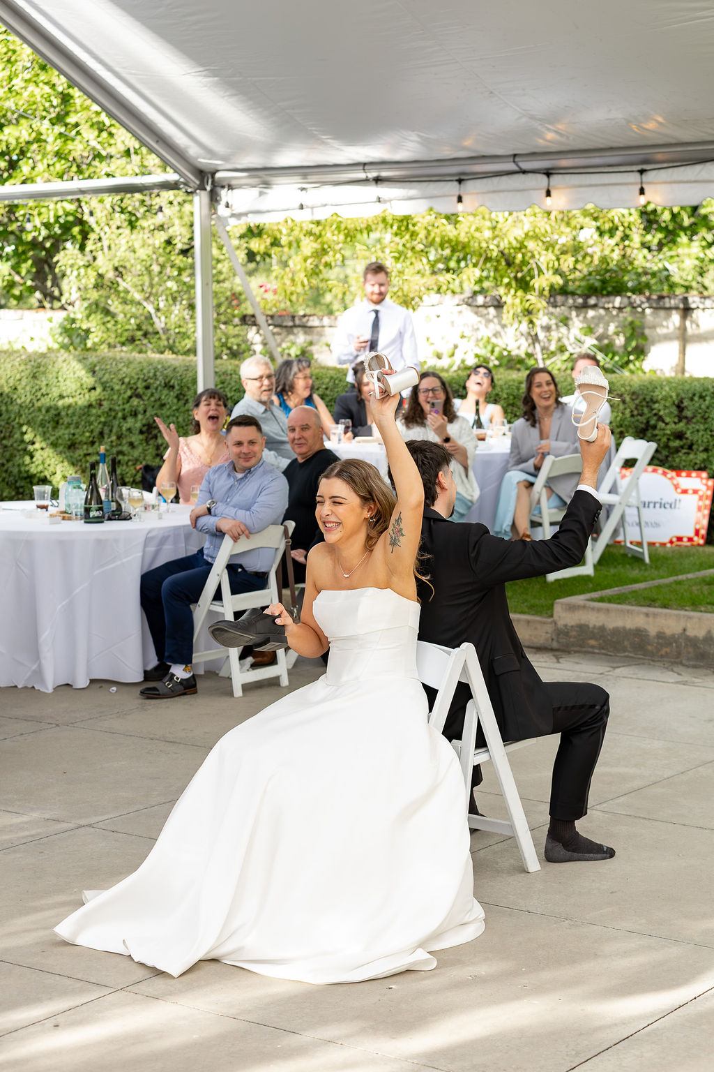 Bride and groom play a game with backs together, holding a cloth, at outdoor wedding reception under a tent, guests watching.
