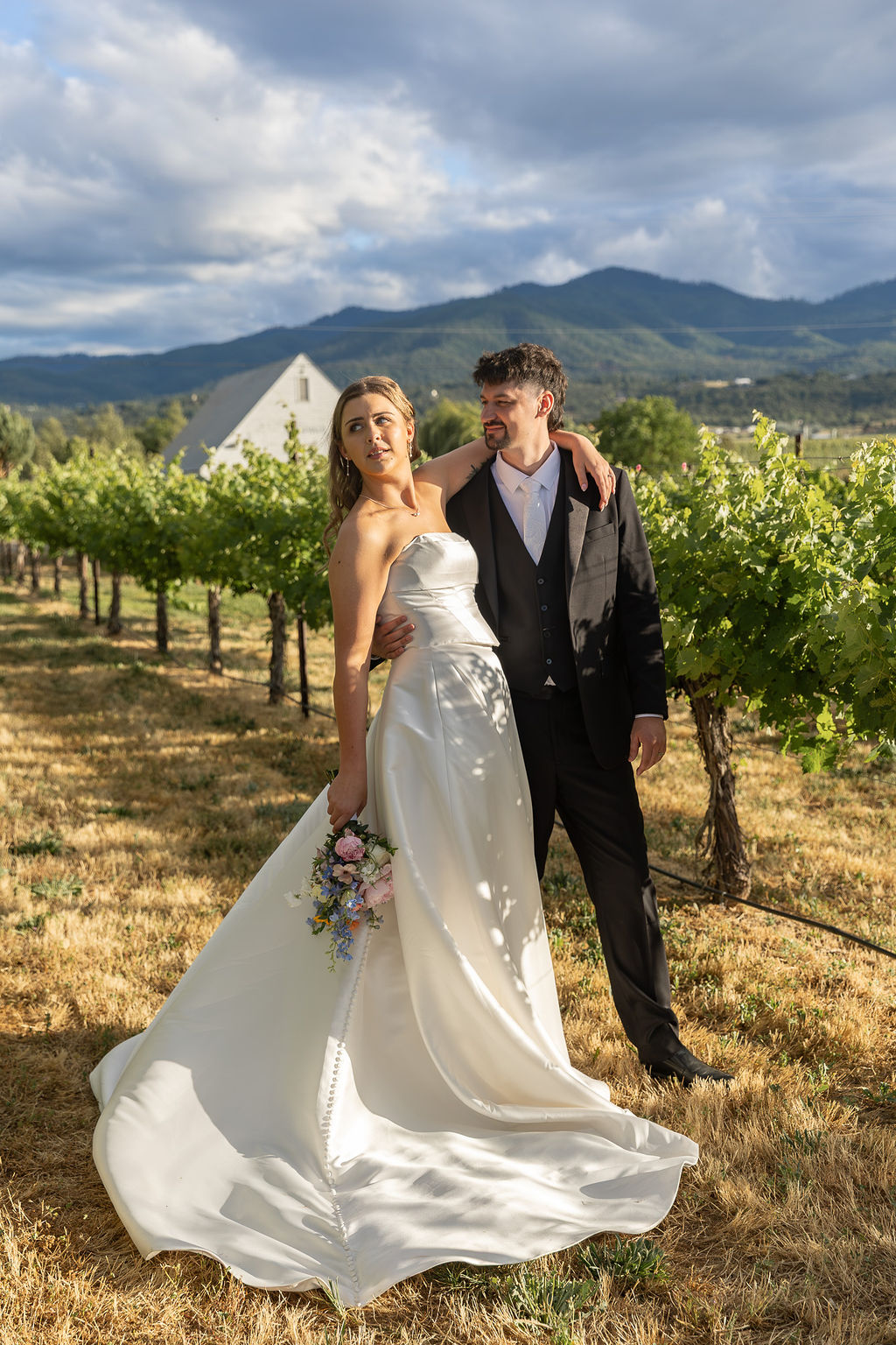 Bride and groom standing in a vineyard with mountains and cloudy sky in background, bride holding a bouquet, wedding attire.