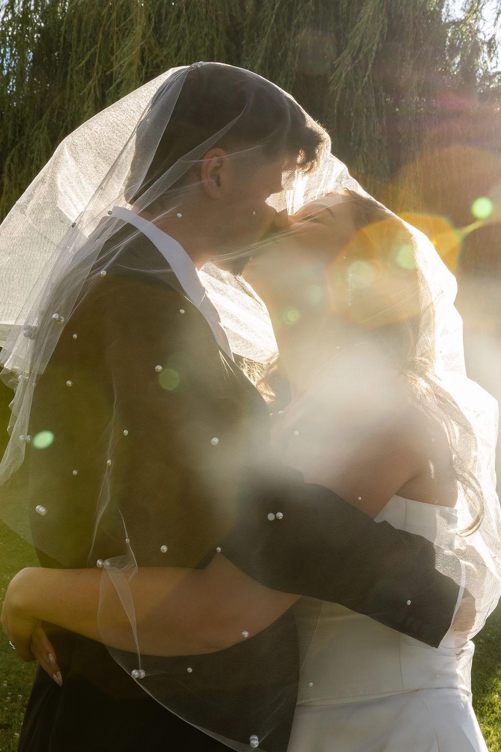 Couple embracing outdoors with sunlight, wearing wedding attire, veil, and smiling, surrounded by greenery.