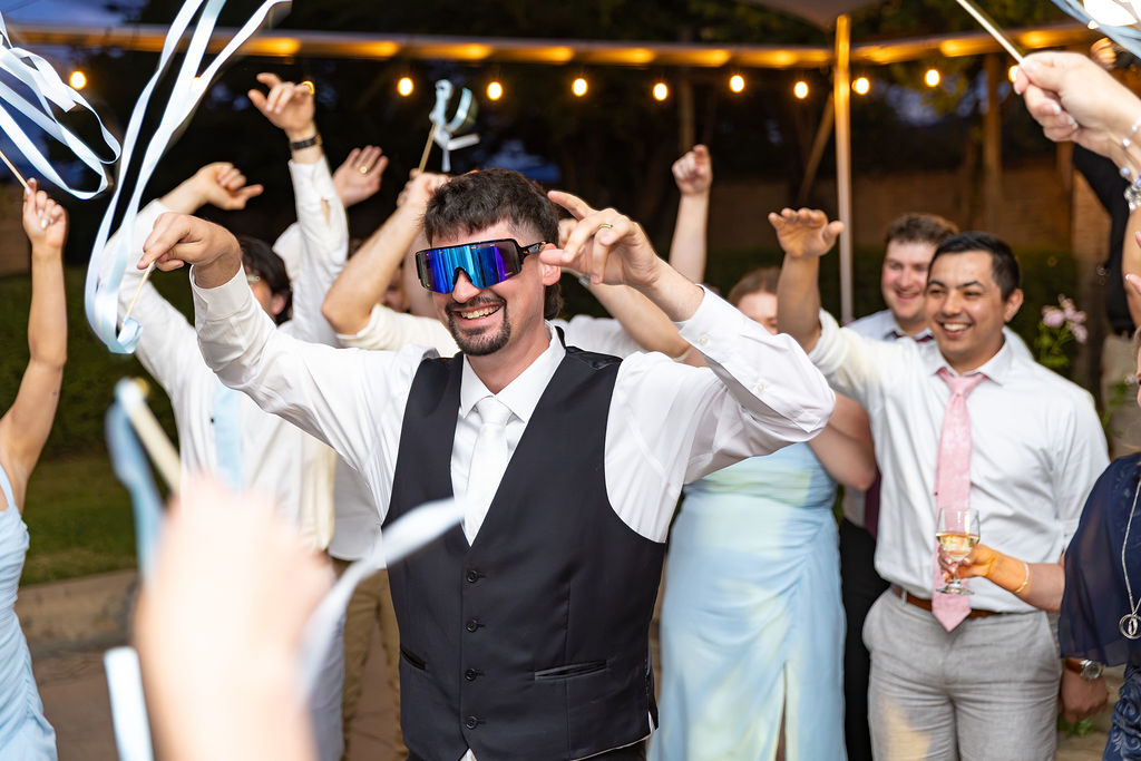 Group of people celebrating outdoors at night, some wearing party accessories, smiling and raising hands, with streamers and lights in background.