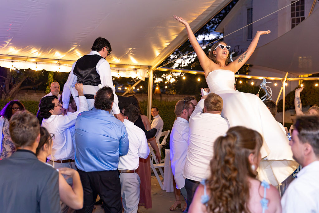 Bride in a white dress and tiara celebrates with guests under a tent at night, some dancing and cheering.