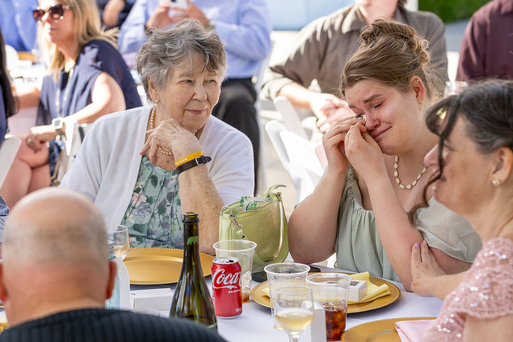 Group of people sitting at a table outdoors, with food and drinks, including a Coca-Cola cup, engaged in conversation.