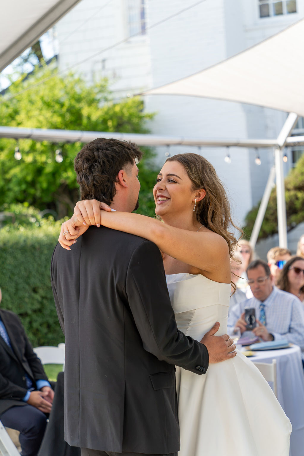 A couple dancing outdoors at a wedding, smiling and embracing, with guests seated at tables in the background.