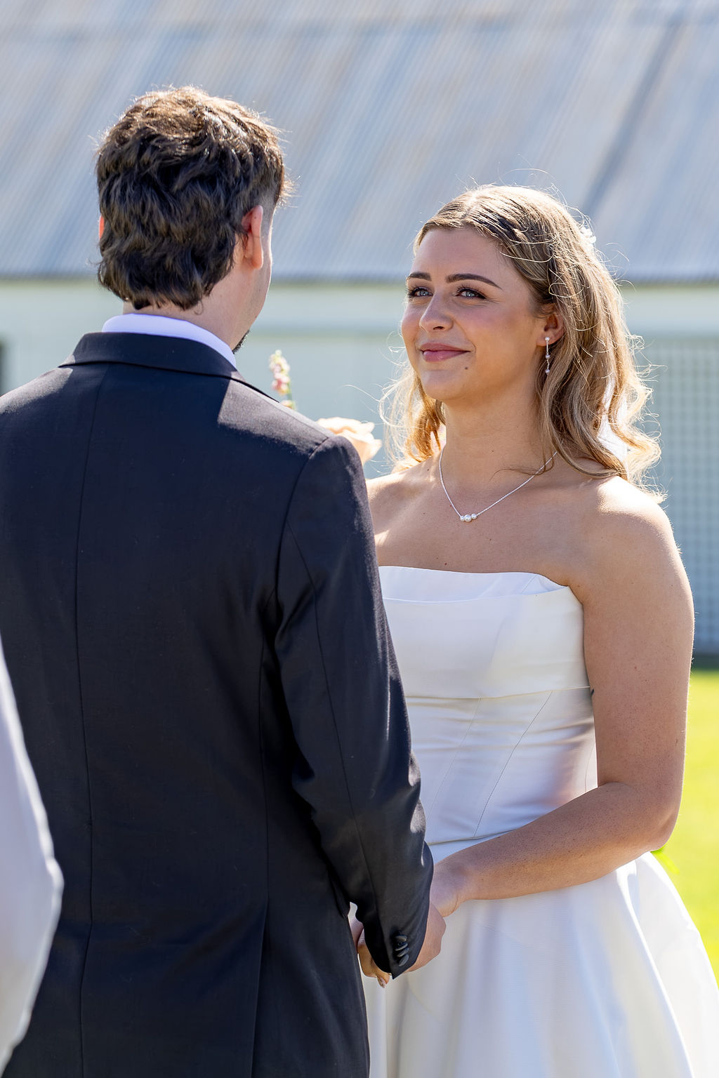 A bride and groom face each other outdoors, with the bride smiling and wearing a white strapless dress.