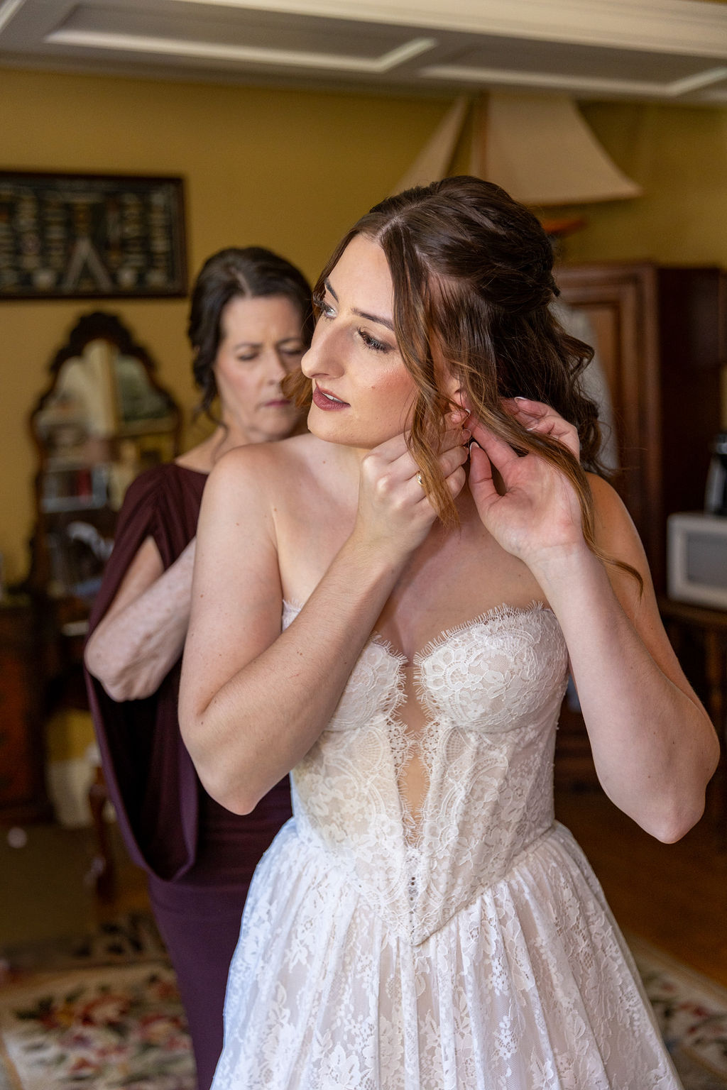 Bride in wedding dress adjusting earring, woman in background helping, indoor setting with warm lighting.