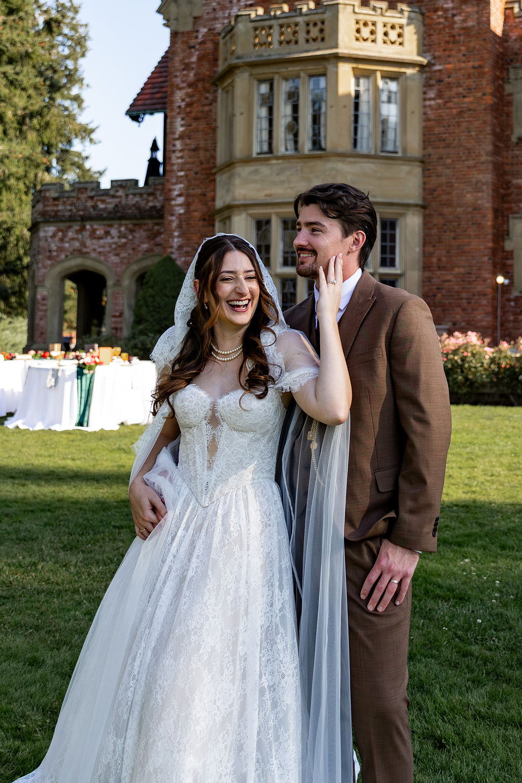 Bride and groom smiling outdoors in front of a historic building, bride in a white wedding dress, groom in a brown suit.