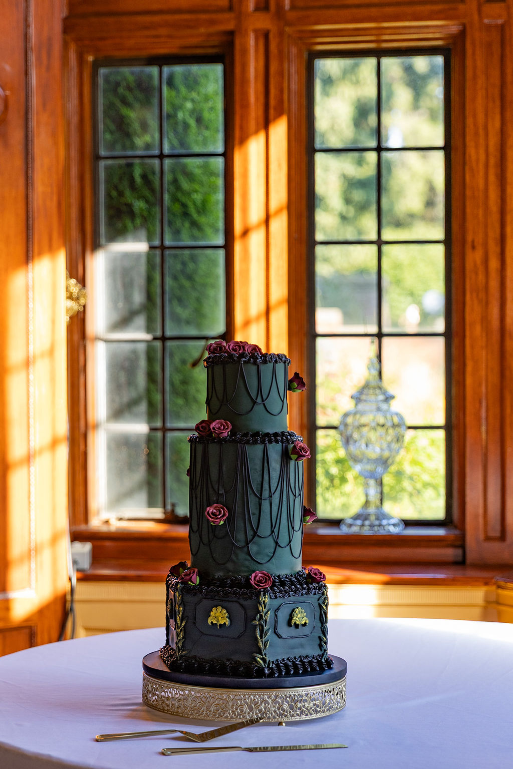 Three-tiered black and purple cake with floral decorations on a table, sunlight streaming through wooden-framed windows, outdoor greenery visible.