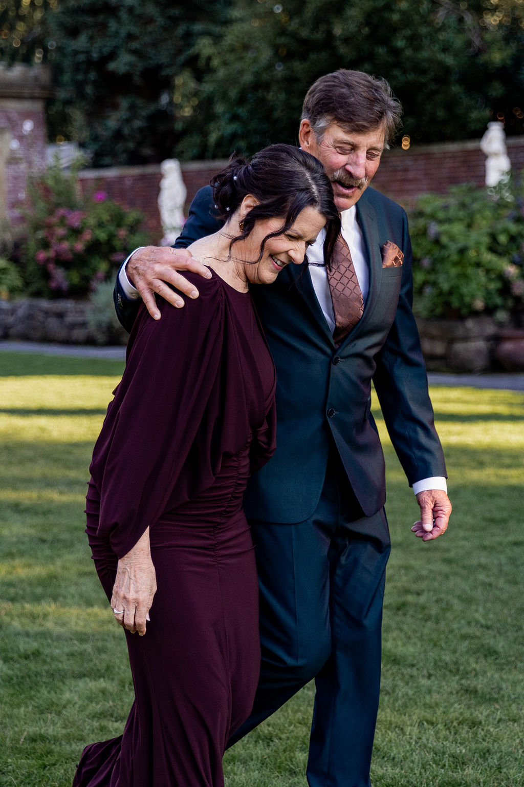 A man and woman smiling and walking outdoors, with the man embracing the woman. They are dressed formally, with greenery in the background.