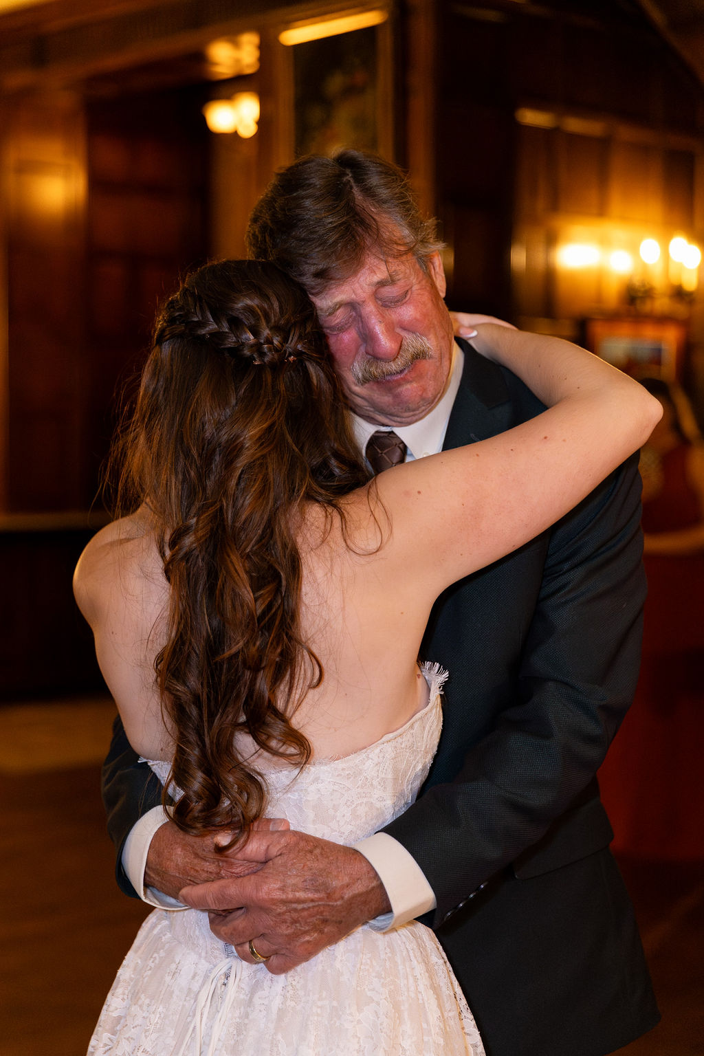 A woman with long hair embraces a man in a suit, both smiling warmly in a warmly lit room.