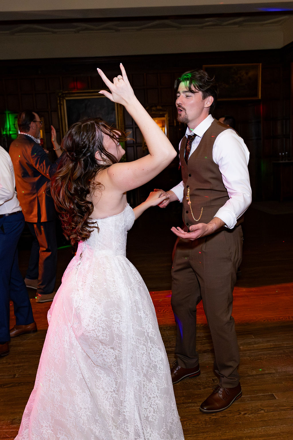 Two people dancing indoors, woman in a white dress and man in a brown vest and white shirt, with other guests in background.