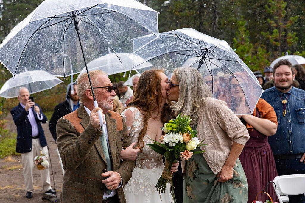 Two women kiss at a wedding, holding a bouquet, with guests and umbrellas in the background outdoors.