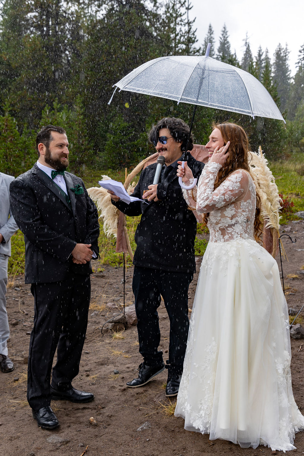 Bride and groom standing outdoors in rain, with officiant holding umbrella, trees in background, wedding attire, and smiling faces.