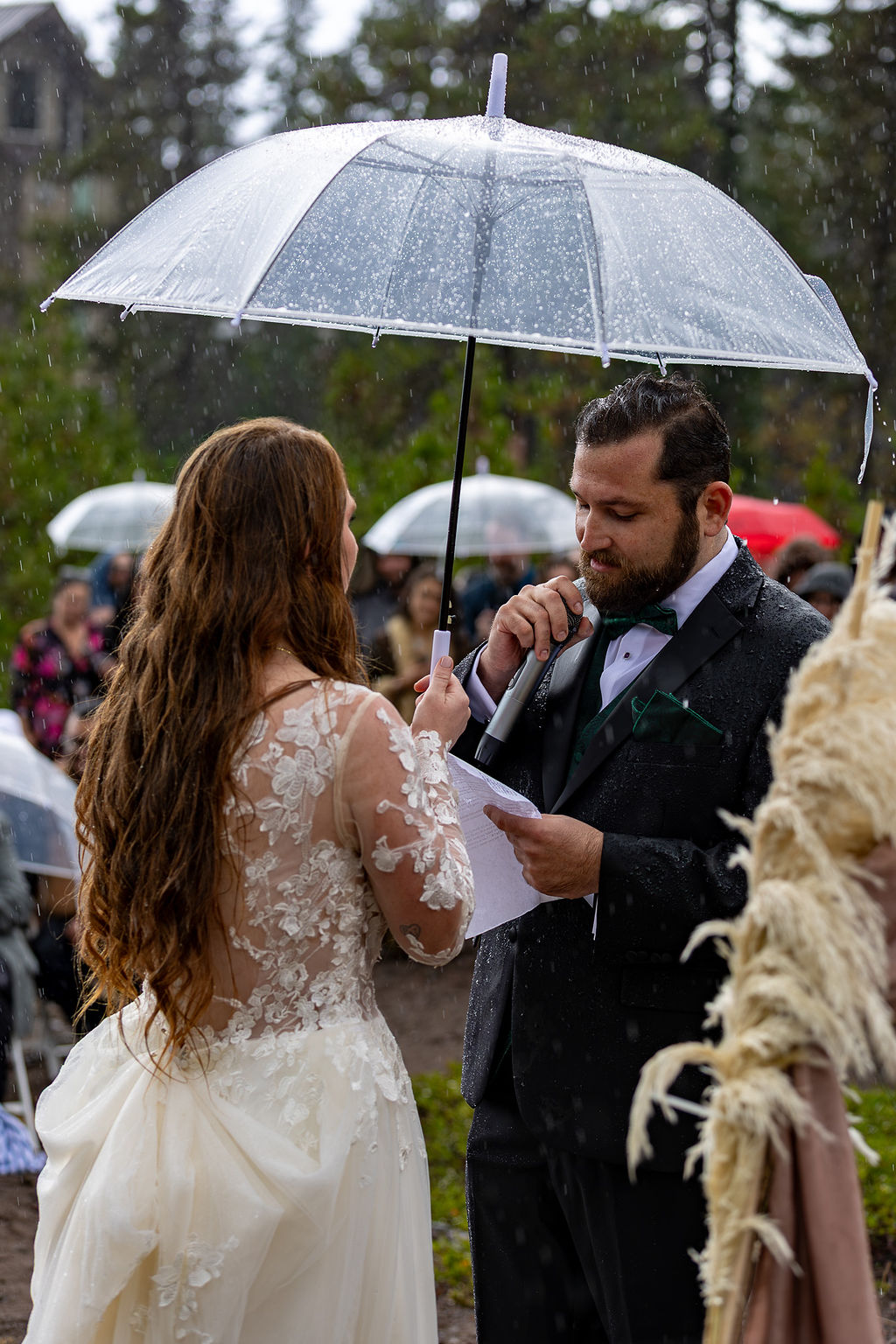 A couple stands under a transparent umbrella during an outdoor wedding ceremony, with guests in the background.
