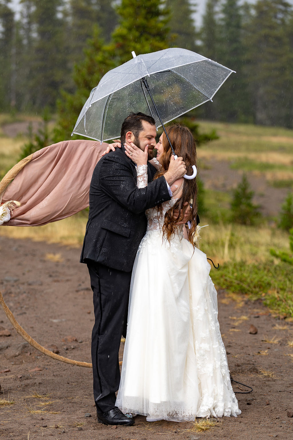 A couple in wedding attire kissing under a transparent umbrella outdoors, with a person lying on a platform behind them.