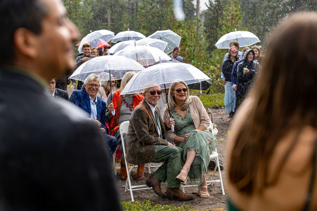People sitting outdoors with umbrellas, some smiling, others watching an event in the rain.