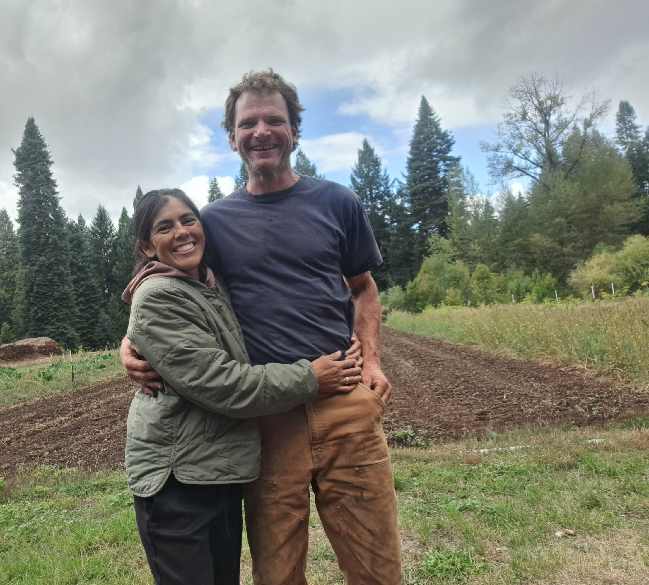 Two people standing outdoors, smiling, with trees and a cloudy sky in the background.