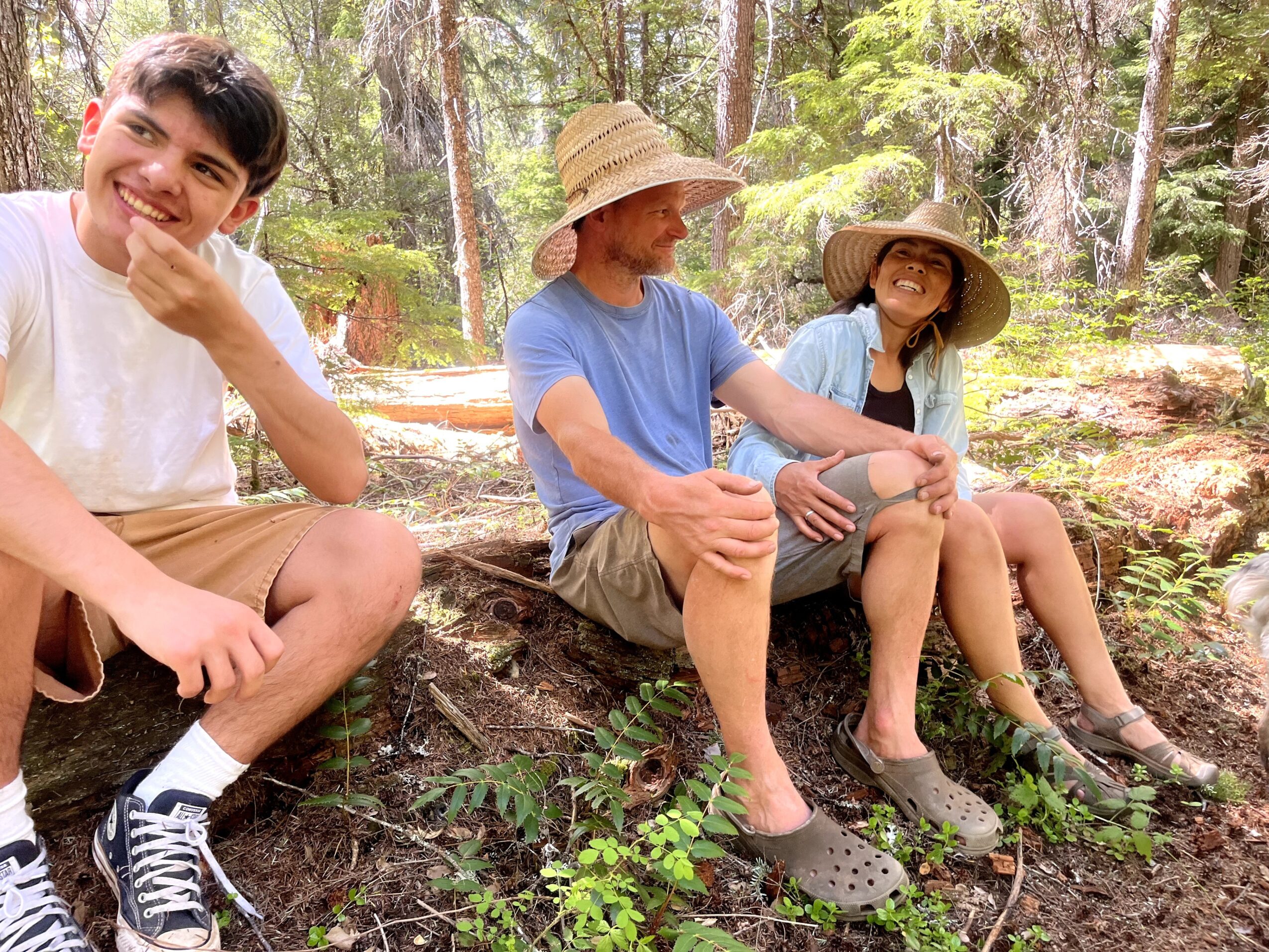 Three people sitting outdoors in a forest, two wearing large straw hats, smiling and relaxing on a log.