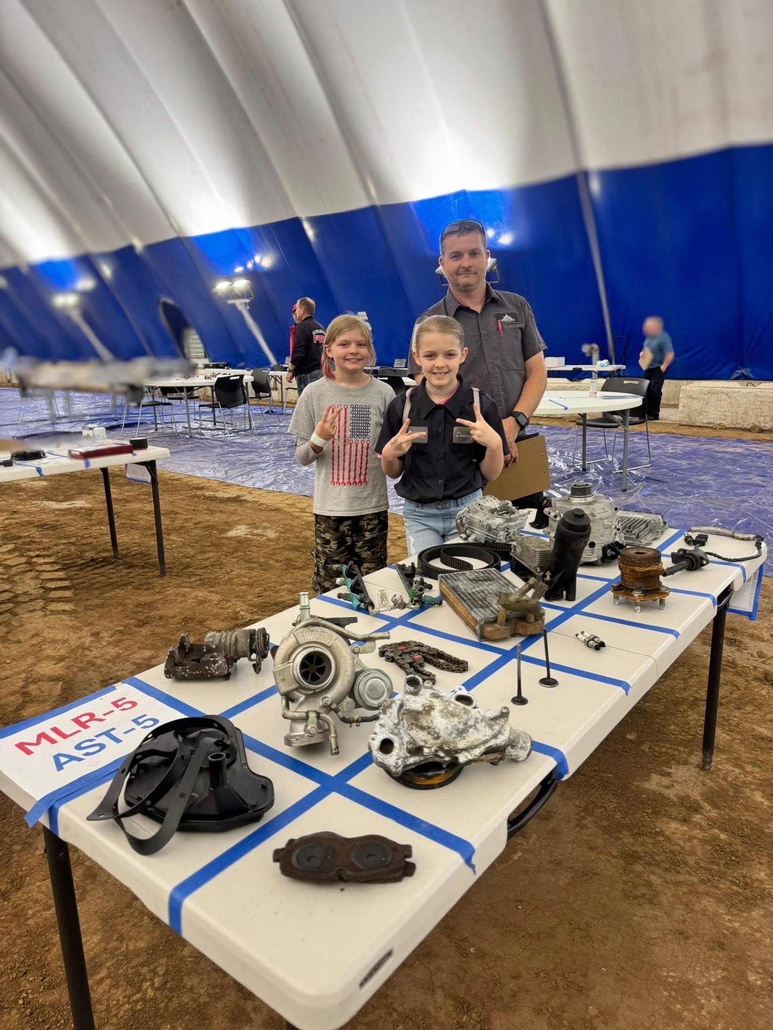 Three people, two children and one adult, stand behind a table with mechanical parts inside a large indoor space with a blue and white wall. Word partially visible on table: 'MLR &'.