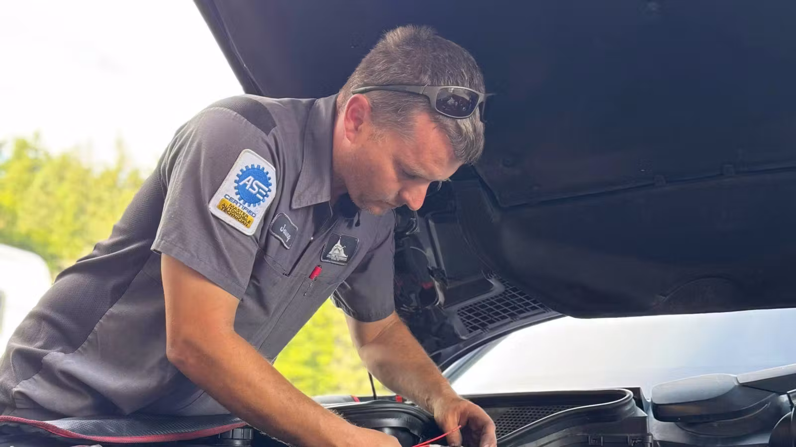 Man in uniform inspecting a car engine with the hood open outdoors.