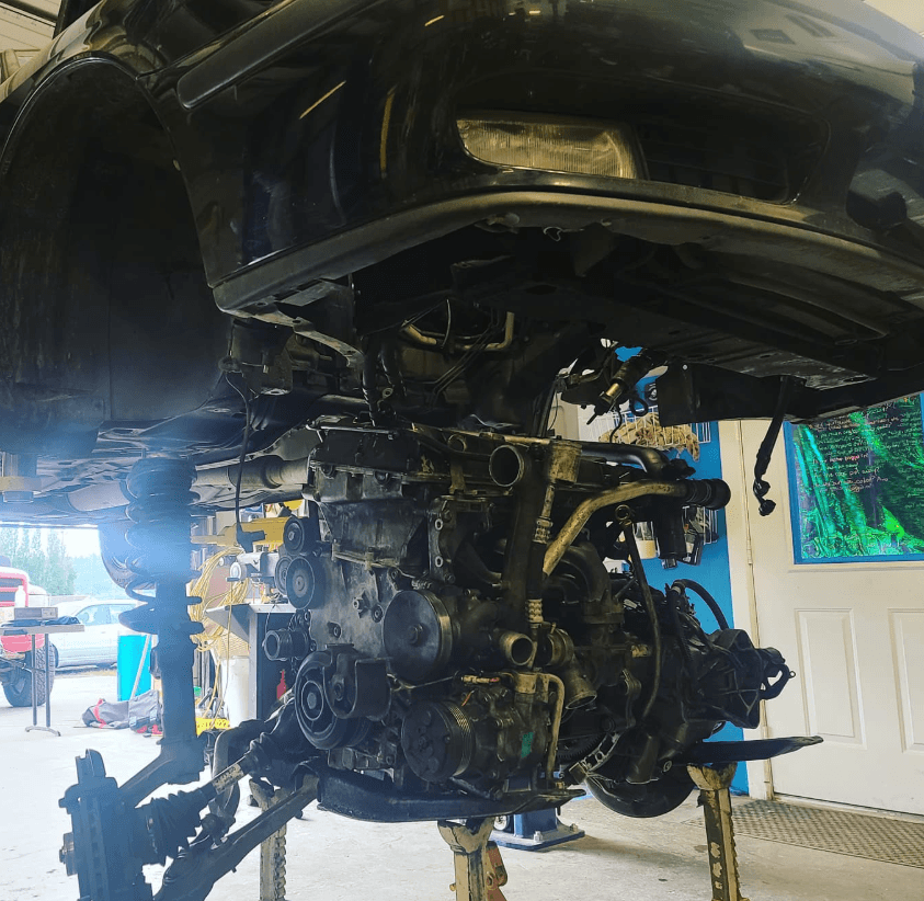 Car engine removed from vehicle, suspended on a lift in a garage, with tools and equipment visible in the background.